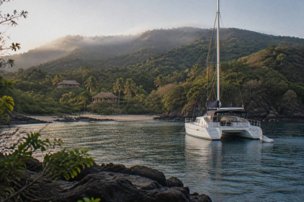 A sailing catamaran moored in a quiet Pacific cove with forested hills behind it
