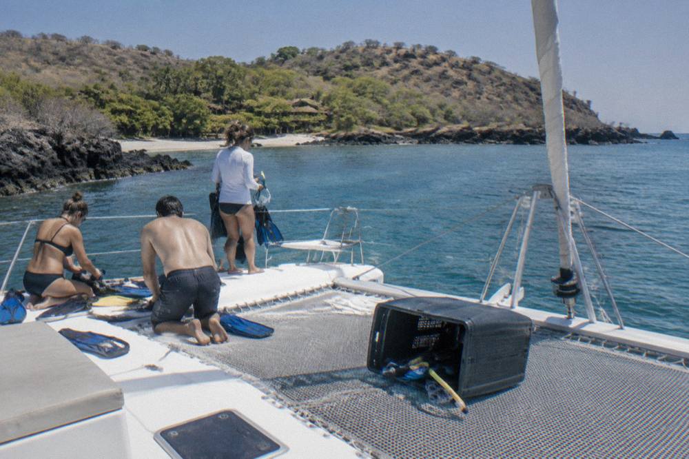 A anchored catamaran with passengers preparing snorkel gear near a rocky shoreline