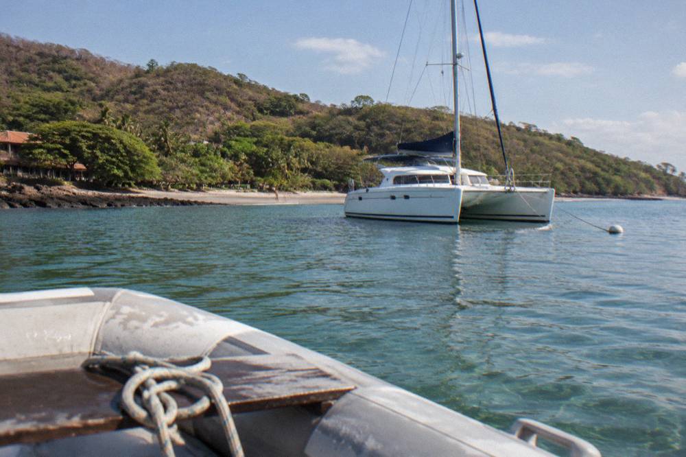 A catamaran moored in clear shallow water beside a forested bay with a dinghy approaching