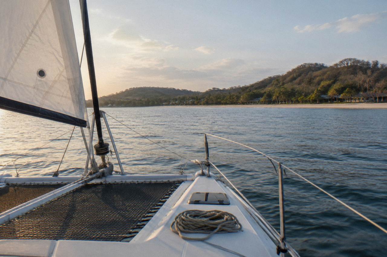 A catamaran under sail on calm Pacific water near a wooded coastline in the late afternoon