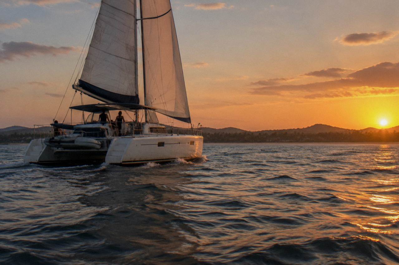 A wide catamaran sailing on calm golden water off a tropical Pacific coastline at sunset