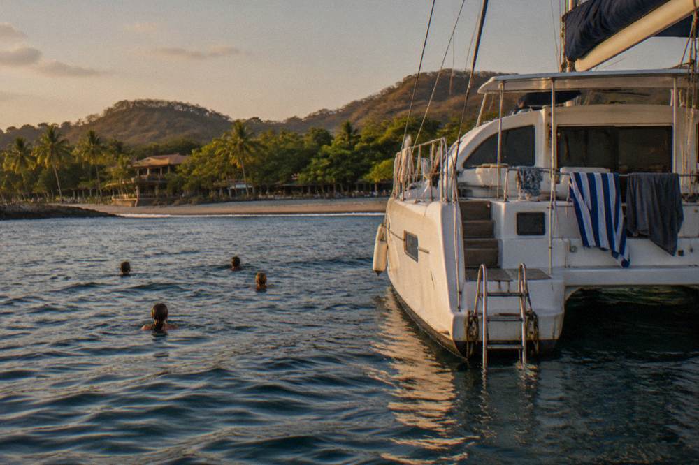 Catamaran anchored in a quiet bay with swimmers in the water beside the hull