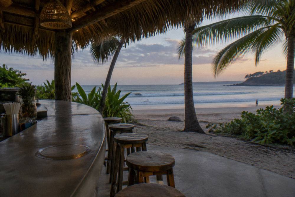 Open-air beach bar with palm-thatch roof and stools facing the ocean at dusk