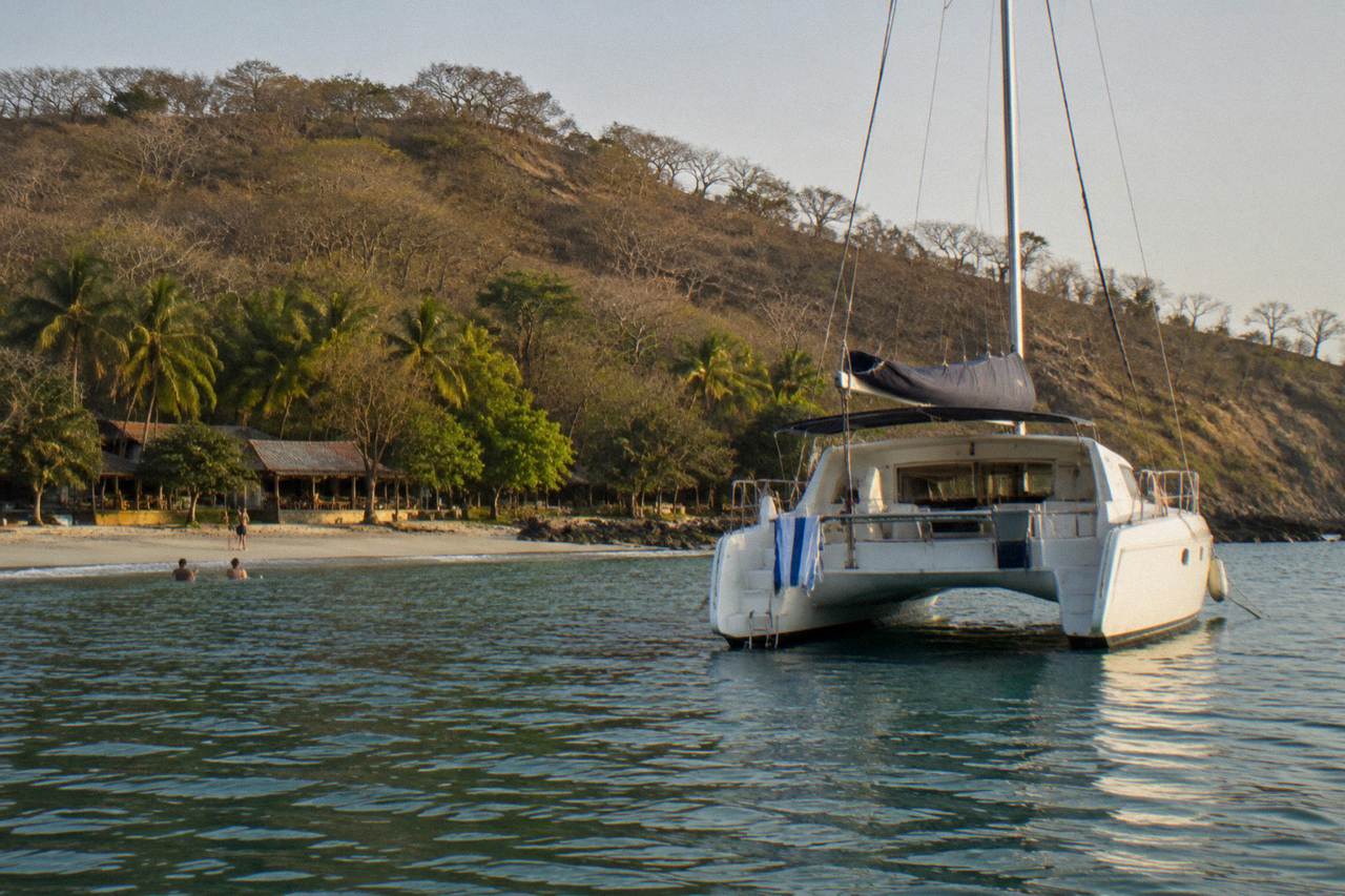 A wide catamaran anchored in calm water near a wooded coastline at late afternoon