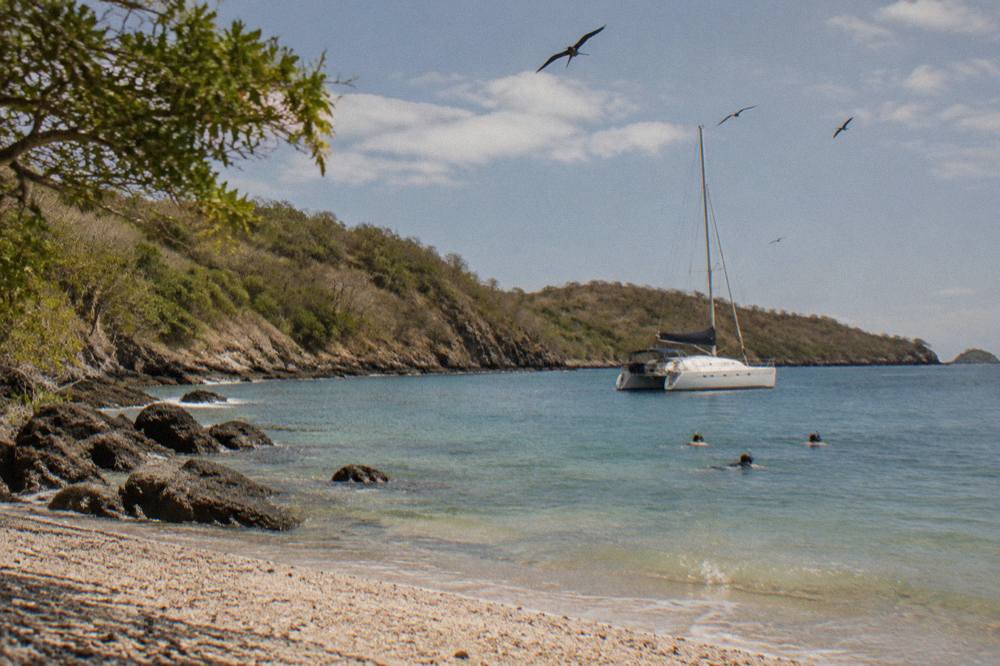 A catamaran anchored in a calm rocky bay with snorkelers in the water near the shore