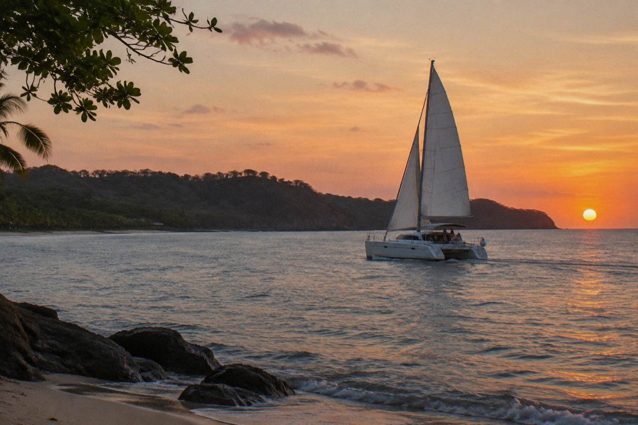 A catamaran under sail on calm Pacific water as the sun drops toward the horizon