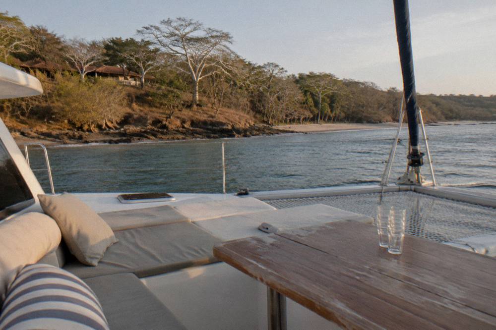 Passenger-free catamaran deck at anchor near a wooded coastline in late afternoon light