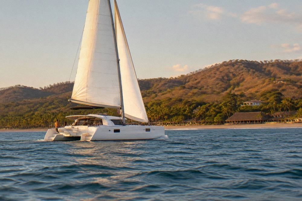 Catamaran under sail on calm Pacific water along a green coastline in late afternoon light