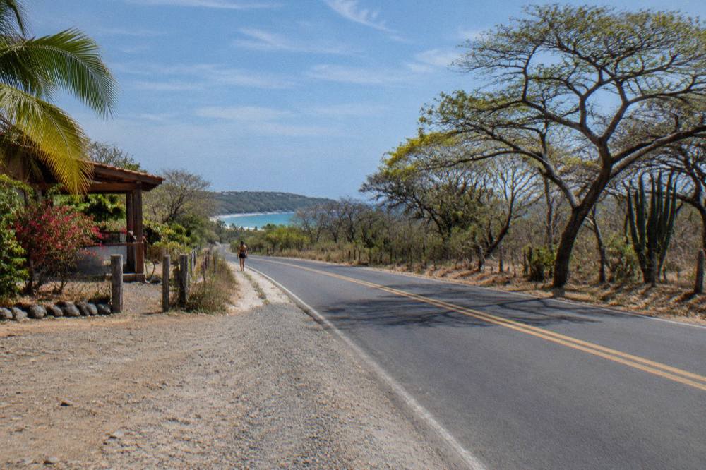 Paved two-lane coastal highway lined with dry forest under a wide sky