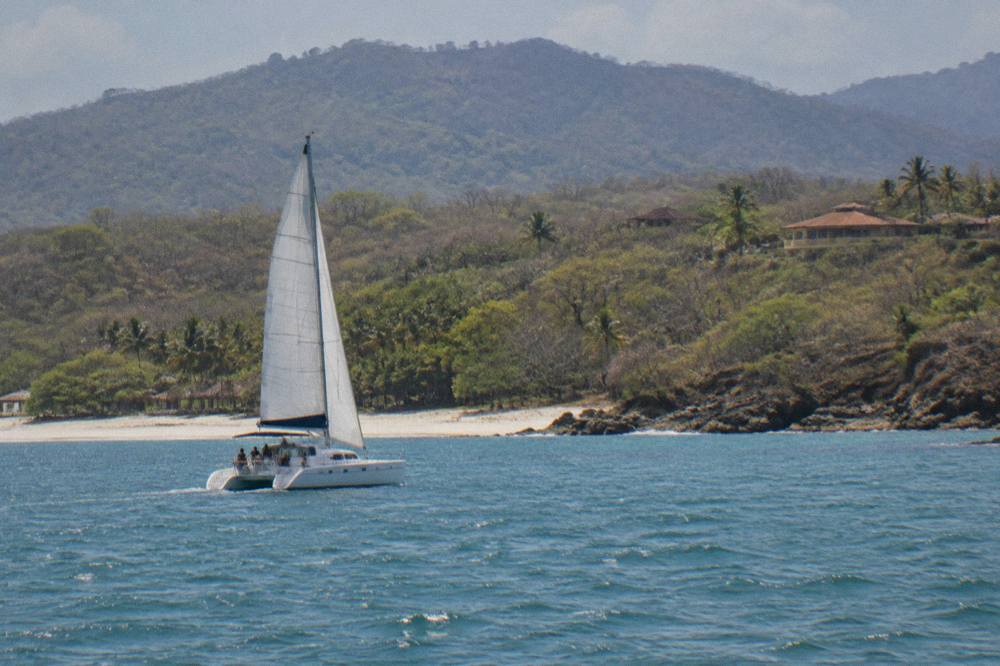 Catamaran sailing along a dry forest coastline toward a pale shell beach