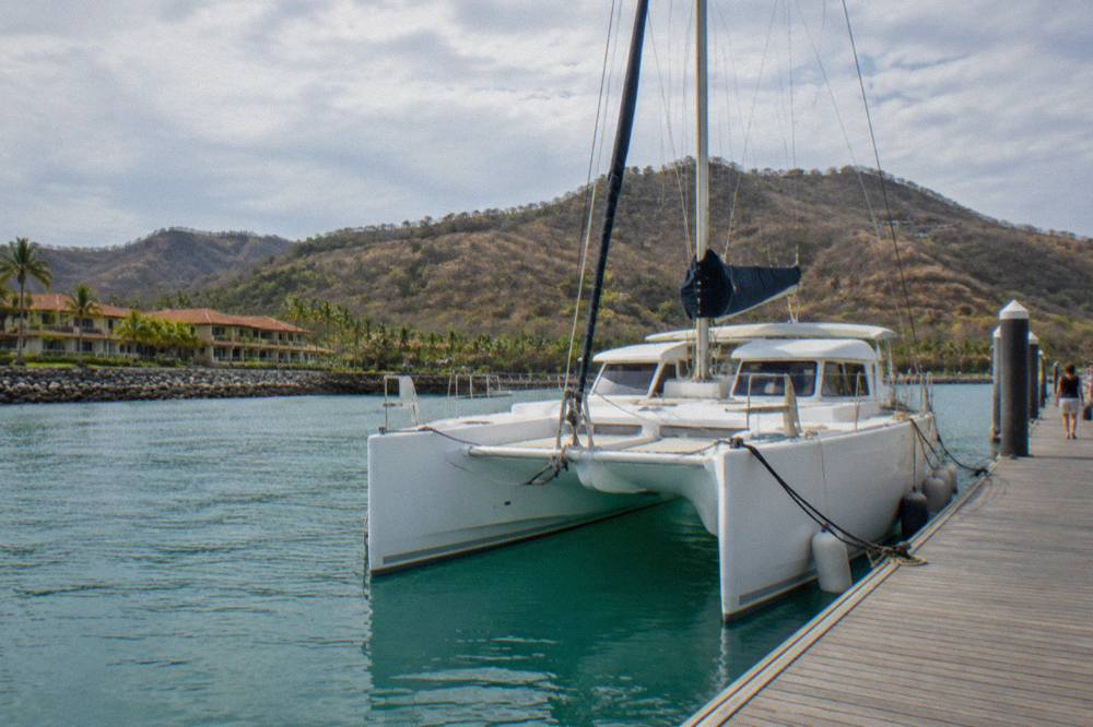 A white catamaran moored at a marina dock with calm turquoise water and wooded hills behind it