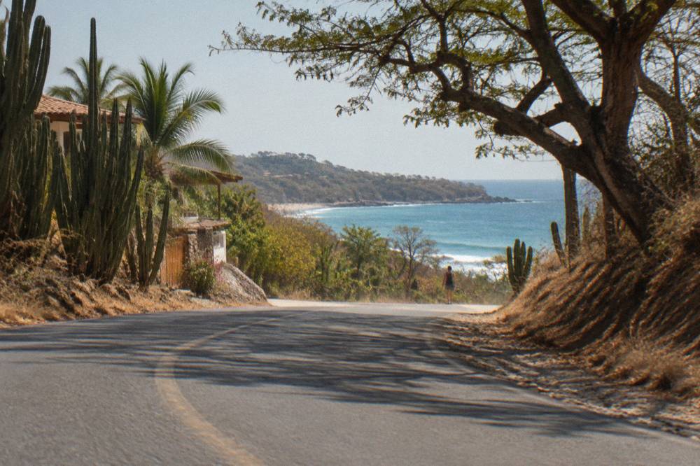 A paved coastal road curving between dry forest hills with the ocean visible ahead