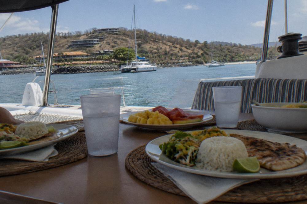 A buffet lunch and drinks arranged on a catamaran deck table under a shaded seating area