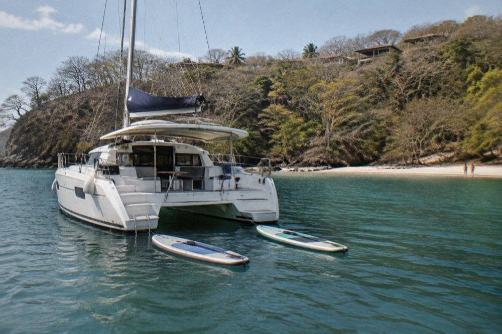 A catamaran anchored in a quiet tree-lined cove with paddleboards floating beside it
