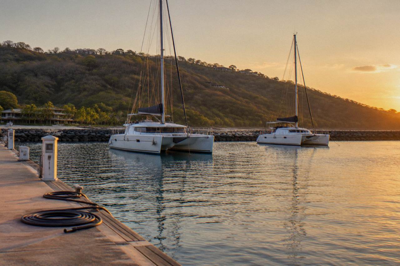 Anchored catamarans resting on calm water beside a marina breakwater at golden hour