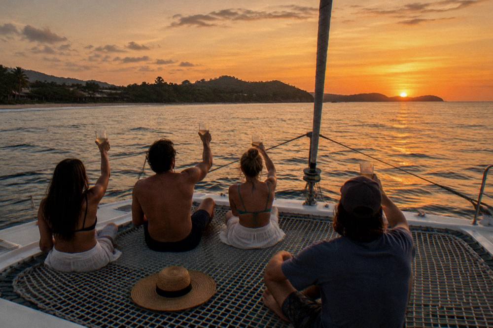 A small group toasting with drinks on the bow of a catamaran near sunset