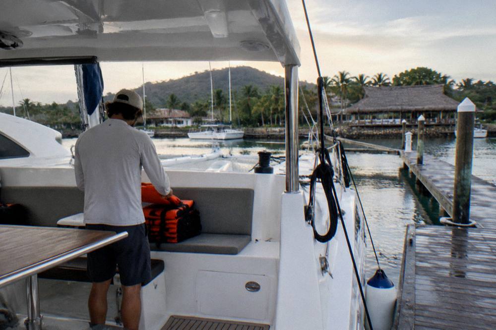 Crew member stowing orange life vests beneath catamaran cockpit seating