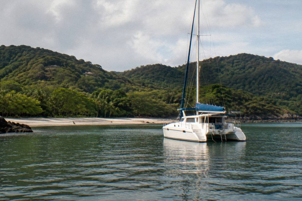 Catamaran at anchor in a quiet green-season bay with forested hills behind