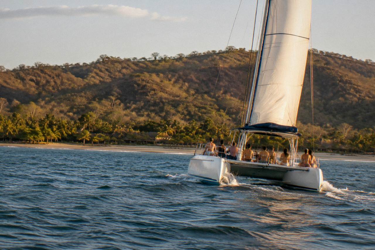Catamaran under sail off a Pacific beach coastline in late afternoon light