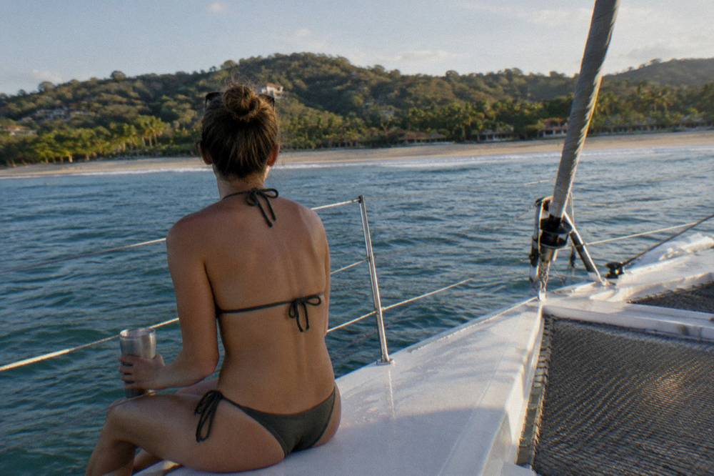 A guest in a swimsuit holding a cold drink at the rail of a catamaran under sail