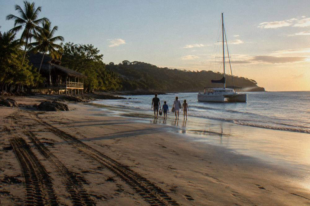 A family walking along a tropical beach toward a catamaran moored offshore