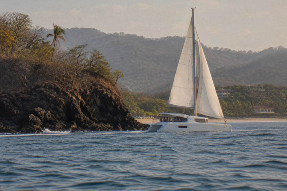 Catamaran with sails up cruising along a rocky Guanacaste shoreline in late afternoon
