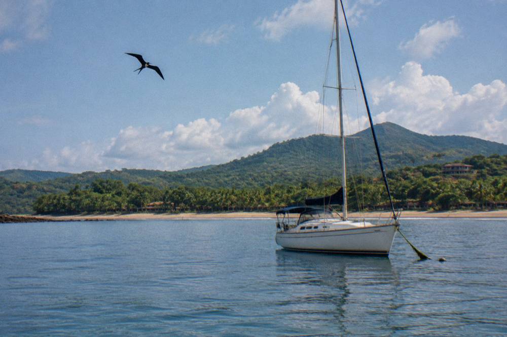 Sailboat at anchor in a calm bay with green forested hills under scattered clouds