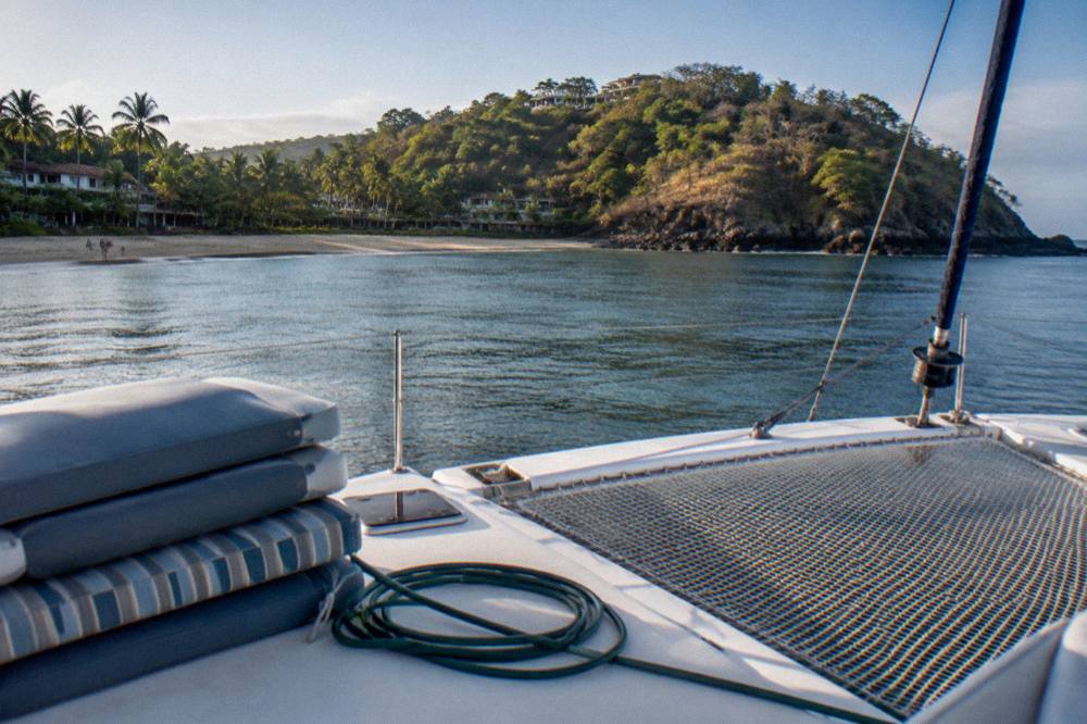 Empty catamaran deck with nets and cushions in bright morning light near a green coastline