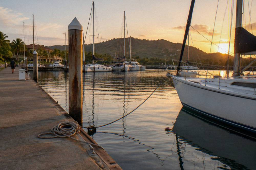 A calm marina at golden hour with several moored sailing boats and green hills behind